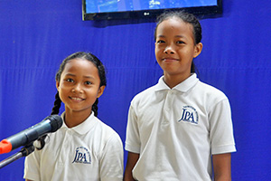 JPA Image Gallery - Two girls smile at the mic on the assembly stage - Jay Pritzker Academy, Siem Reap, Cambodia