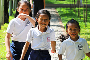 JPA Image Gallery - Three primary students laugh as they make their way down a path on campus - Jay Pritzker Academy, Siem Reap, Cambodia