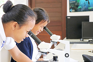 JPA Image Gallery - 2 students examine something with a microscope - Jay Pritzker Academy, Siem Reap, Cambodia