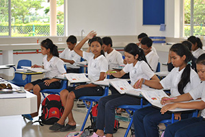JPA Image Gallery - A student raises her hand to ask a question while reading a textbook in class - Jay Pritzker Academy, Siem Reap, Cambodia