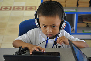 JPA Image Gallery - A young primary student wearing headphones focuses on the computer in front of him - Jay Pritzker Academy, Siem Reap, Cambodia